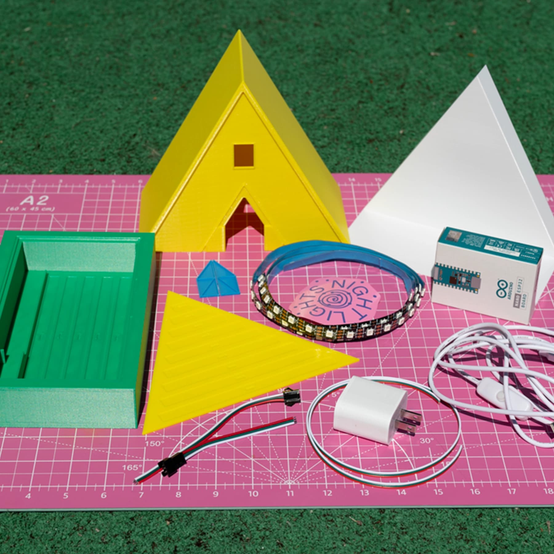 The Temple DIY Kit contents on a pink cutting mat: yellow 3D-printed A-frame shell, white inner light diffuser, green base, small accent pieces, an addressable LED strip, an Arduino Nano, a USB cable with inline switch, a wall adapter, and connector wires.