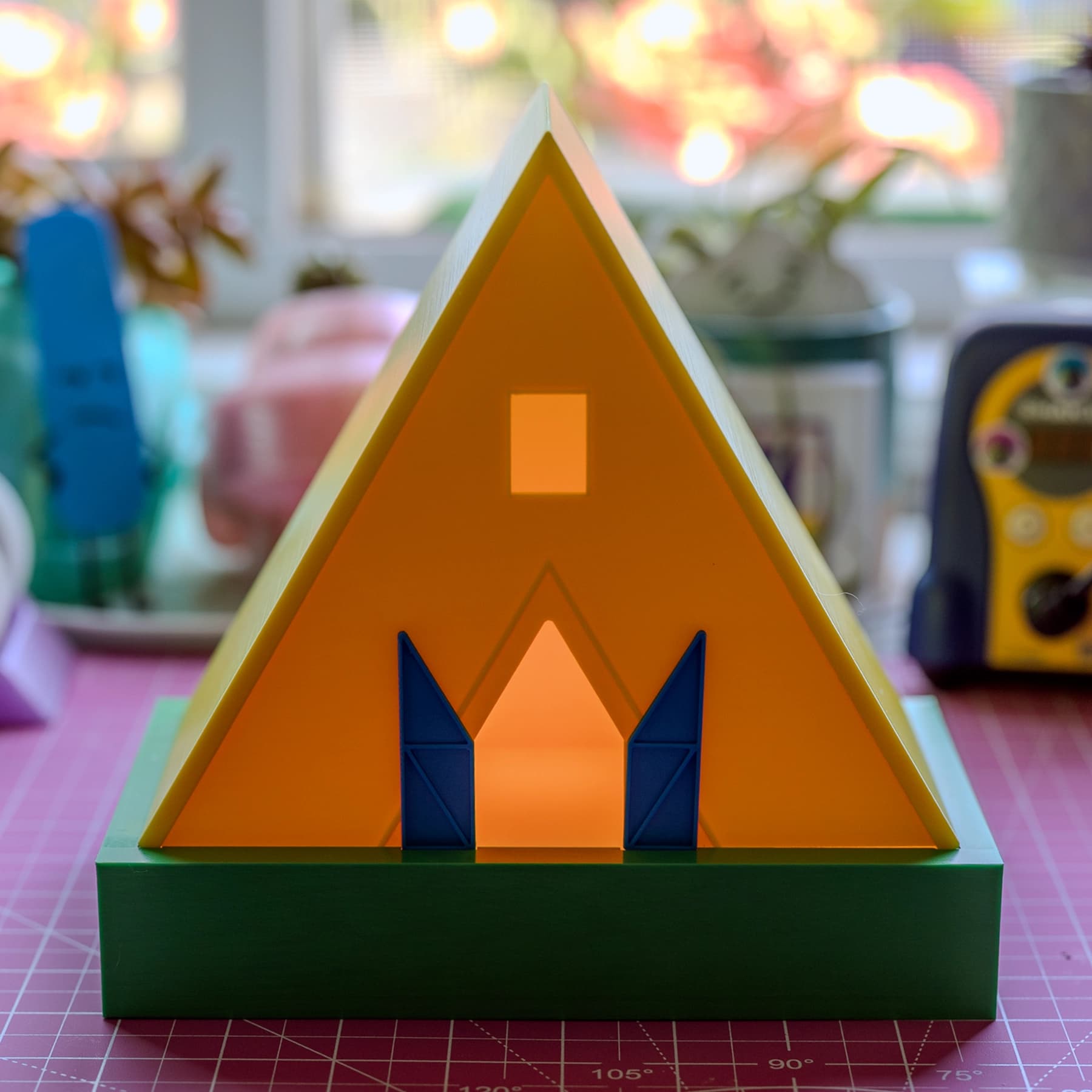 A yellow 3D-printed A-frame temple lamp on a solid green base, its orange front face glowing warm, with two blue doors flanking a lit doorway and a small square window near the peak.